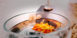 Eine orangefarbene Blume schwimmt in einer Glasschale mit Wasser, im Hintergrund unscharfe Objekte.