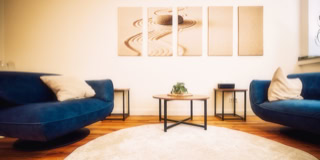 Ein Wohnzimmer mit zwei blauen Sofas, einem runden wei&szlig;en Teppich, einem kleinen Holztisch mit Pflanze und vier abstrakten Bildern an der Wand.