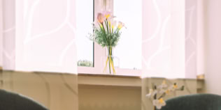 Ein Blumenstrau&szlig; in einer Vase steht auf einer Fensterbank vor einem Fenster mit wei&szlig;en Vorh&auml;ngen.